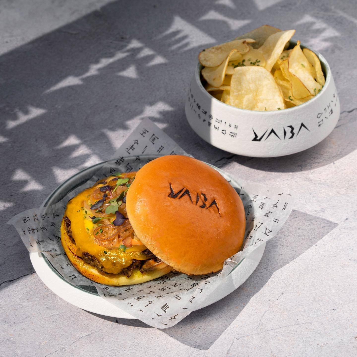 The YABA wordmark branded onto a burger bun, served on cuneiform-printed paper beside a matching bowl